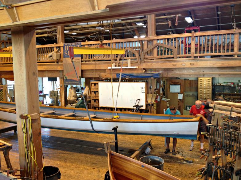 wooden workshop with tools and a large rowing gig suspended from a gantry lift with three men looking at the boat's bow