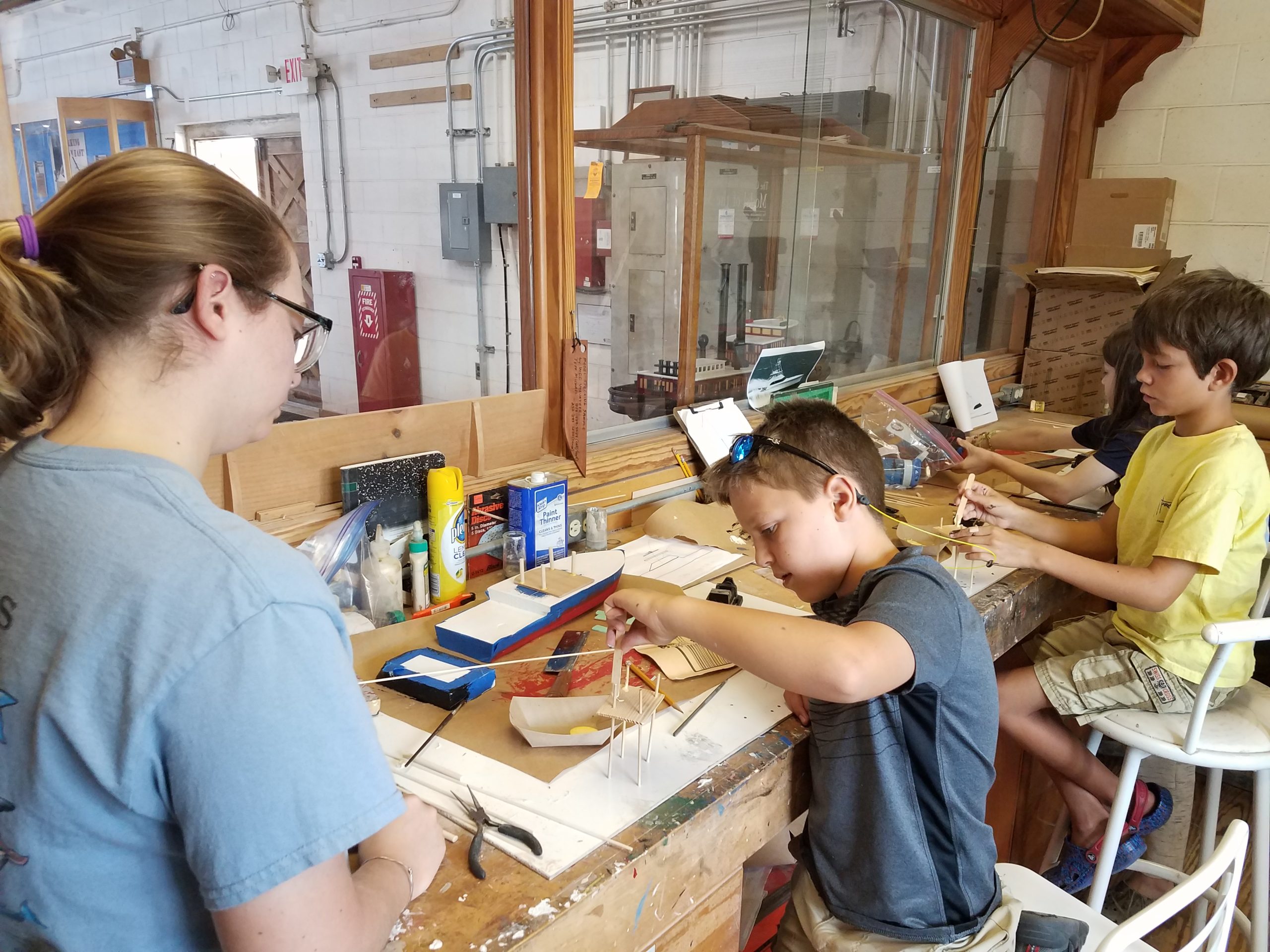 two boys sit at a bench working on wooden boat models as an adult watches