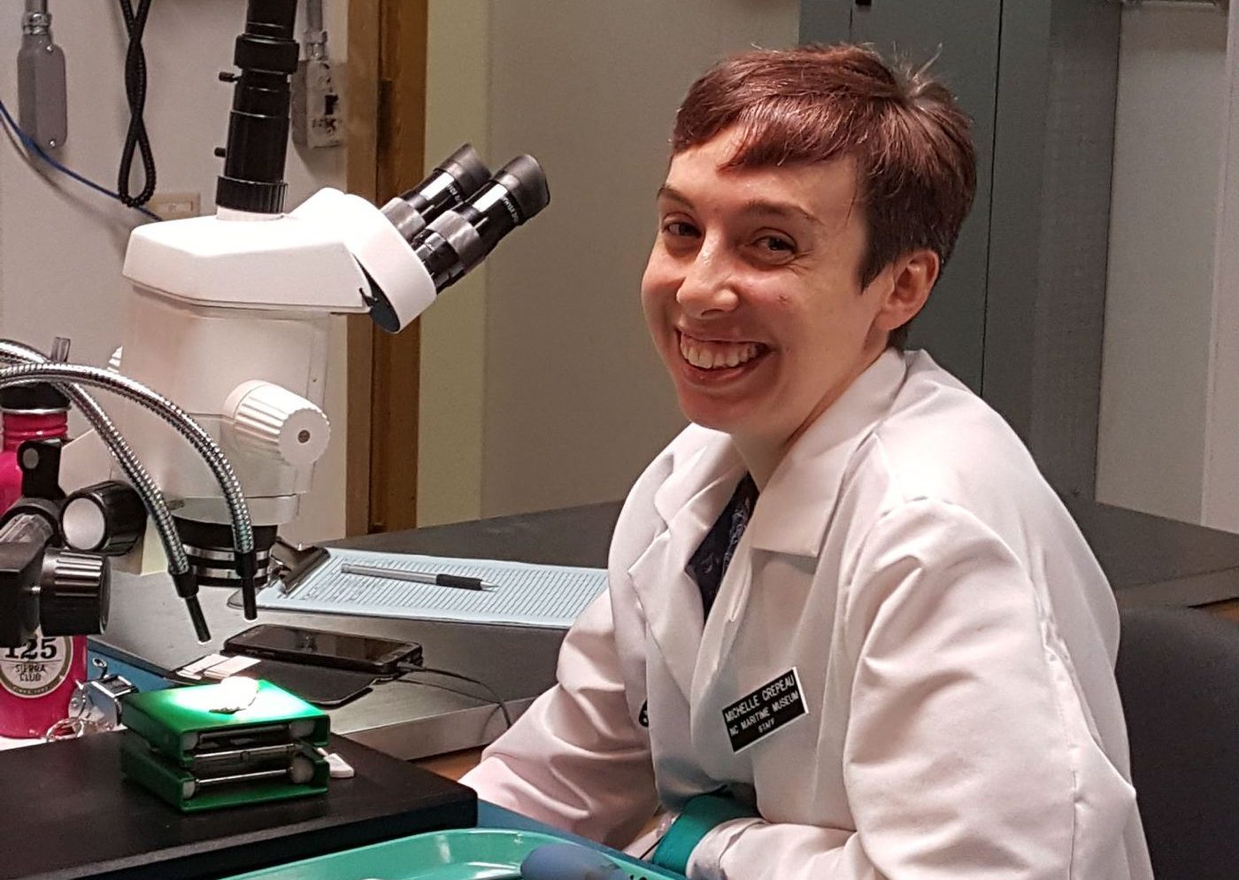 woman in lab coat next to microscope looking at camera and smiling