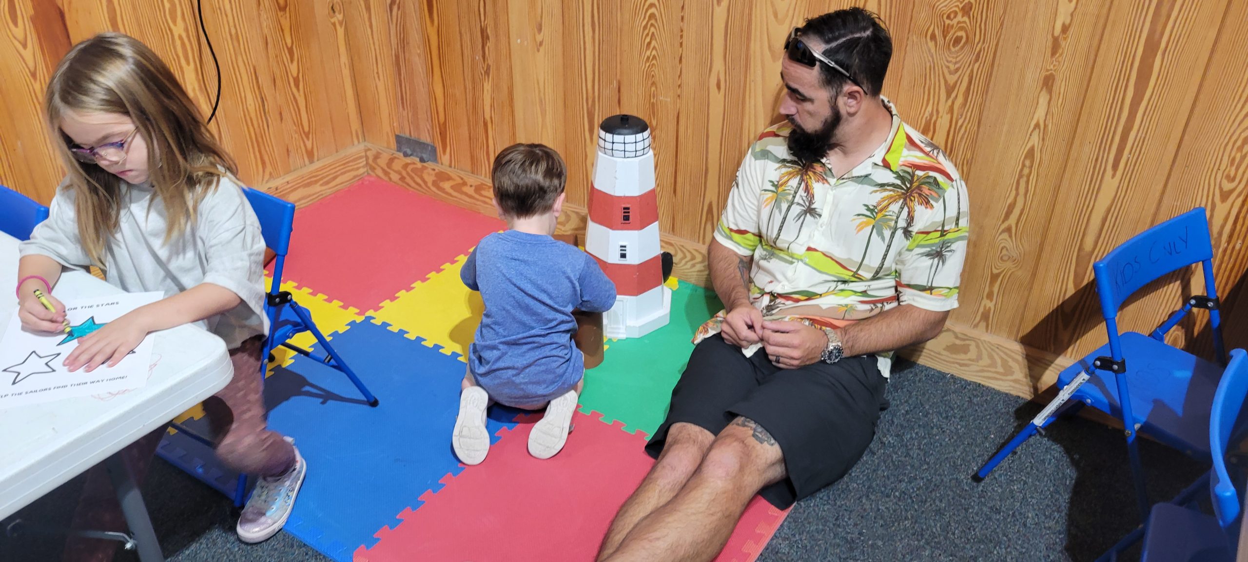 man sitting with back against wall watches a young boy play with a puzzle while a young girl sits coloring at a table nearby