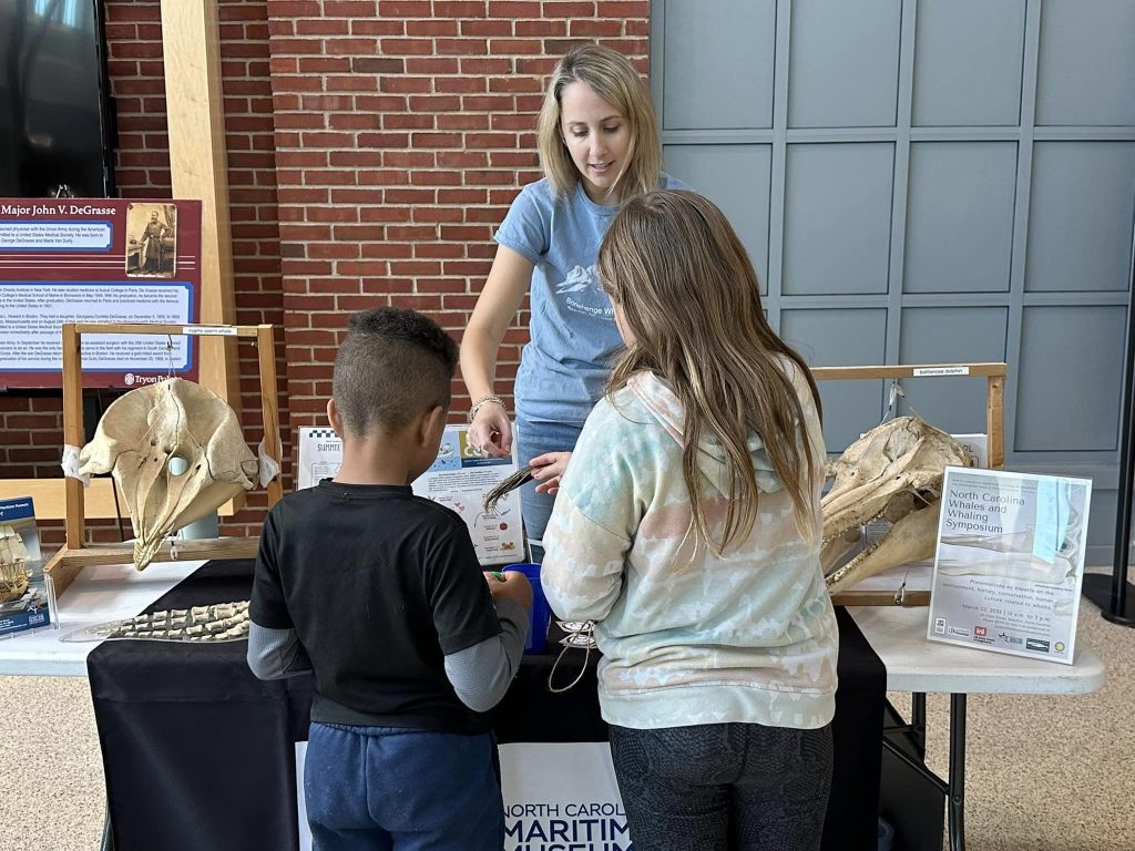Woman stands at display table with two children facing the table and exploring the items on it